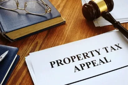 A close-up image of a document titled "PROPERTY TAX APPEAL" on a wooden table with a gavel and a pair of eyeglasses on an open book in the background.